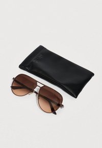 Καφέ φιμέ γυαλιά ηλίου με μεταλλικό πλαίσιο, με γεωμετρικό σχέδιο και μαξιλαράκια στη μύτη, συνοδευόμενα από ένα μαύρο δερμάτινο θήκη με ανάγλυφο το λογότυπο "QUAY".