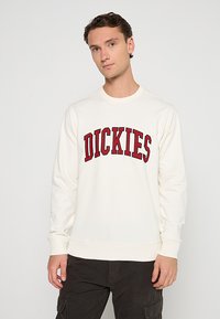 Felpa color crema con maniche lunghe e scollo a girocollo lavorato a maglia. Logo "DICKIES" ricamato in rosso sul petto. Tessuto misto cotone.
