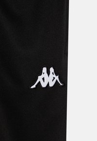 Zwarte stof met een in het wit geborduurd Kappa-logo, dat twee zittende figuren weergeeft, met een zachte textuur en contrasterende kleuren.