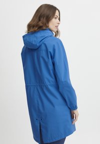 Blå vattentät jacka med huva, elastiska ärmslut och justerbar midja. Har en ventilerad nederkant och en slät tygtextur.