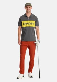 Grijze poloshirt met een felgele horizontale streep en zwart logo, gecombineerd met oranje broek. Het model houdt een golfclub vast en draagt een pet.