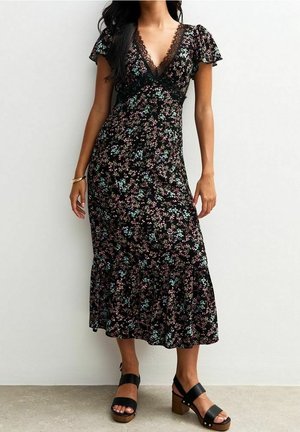 Černé květinové maxi šaty s krátkými volánkovými rukávy, krajkovým lemováním u výstřihu a rozšířeným lemem. Nošeny s černými sandály s robustním podpatkem.