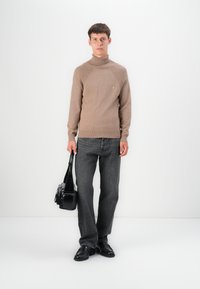 Pull à col roulé en tricot marron clair avec des détails côtelés aux épaules, associé à un jean gris foncé et un sac à bandoulière noir. Chaussures noires.