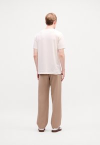Personne se tenant de dos, portant une chemise blanche ample à manches courtes, un pantalon large beige, des chaussettes blanches et des sandales marron, sur fond blanc.