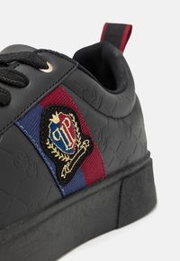 Black sneaker with textured material, featuring a burgundy and blue stripe and a decorative logo patch. Rounded toe and flat rubber sole.
