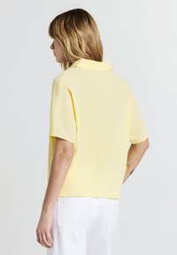 Donna con capelli biondi che indossa una camicia corta di un giallo chiaro con texture e pantaloni bianchi, rivolta verso sinistra su uno sfondo chiaro e semplice.