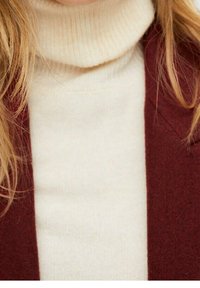 Pull à col roulé crème avec une texture côtelée sous un manteau bordeaux. Gros plan sur le col et les épaules, mettant en valeur le contraste des couleurs.