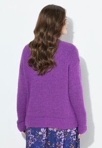 Frau mit langen welligen braunen Haaren, die einen lila gestrickten Pullover und einen blauen Blumenrock trägt, steht vor einer weißen Wand und ist leicht nach rechts gerichtet.