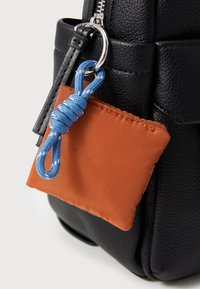 Μαύρη υφασμάτινη τσάντα με μια μικρή πορτοκαλί θήκη προσαρτημένη με μπλε κορδόνι. Η θήκη έχει λεία επιφάνεια και τα αξεσουάρ είναι ασημένια.