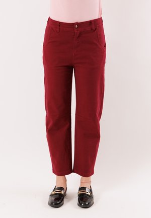 Persona che indossa pantaloni rossi scuro a taglio dritto, mocassini neri con dettagli dorati, in piedi contro uno sfondo chiaro e semplice.