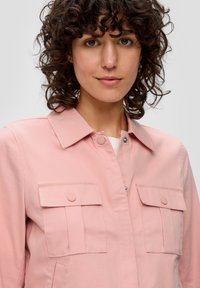 Helle rosa Jacke mit durchgehender Knopfleiste, zwei Brusttaschen und klassischem Kragen. Aus glattem Stoff mit leichtem Glanz gefertigt.