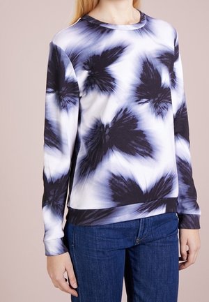 Sweatshirt met lange mouwen in tie-dye-stijl met zwart-witte uitbarstingspatronen, gedragen met een blauwe spijkerbroek door een staande persoon.