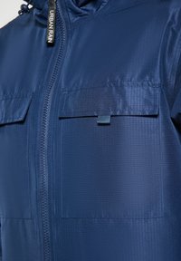 Jūros paviršiaus lietaus striukė su tekstūruotu audiniu, priekinis užtrauktukas, dvi krūtinės kišenės ir reguliuojama kepurė. Prie užtrauktuko yra Urban Rain prekės ženklo logotipas.