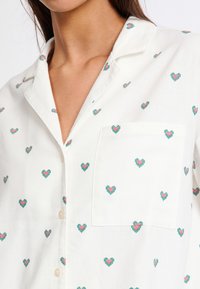 Weißes Hemd mit kleinen, bunten Herzen (rosa und grün) und einer einzelnen Brusttasche; hat einen klassischen Kragen und eine durchgehende Knopfleiste.