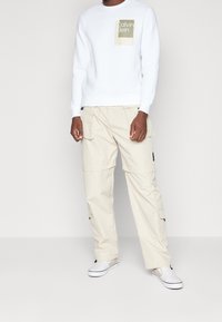Sudadera blanca de cuello redondo con un logo cuadrado verde oliva. Pantalones cargo beige con bolsillos y correas ajustables. Persona vistiendo zapatillas blancas sin cordones.