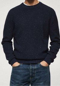 Mężczyzna w granatowym swetrze z wełny z drobnym wzorem, z dłońmi w kieszeniach niebieskich dżinsów, na jednolitym tle.