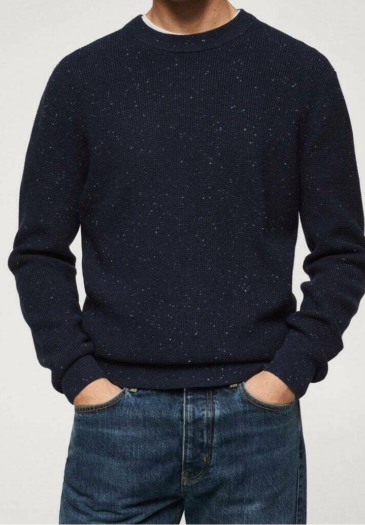 Mężczyzna w granatowym swetrze z wełny z drobnym wzorem, z dłońmi w kieszeniach niebieskich dżinsów, na jednolitym tle.