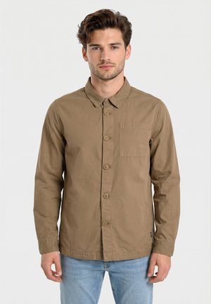 Man met een bruin overhemd met knopen en een lange mouw en lichtblauwe spijkerbroek staat tegen een effen witte achtergrond en kijkt recht vooruit.