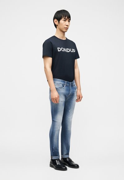 Νέος άνδρας που φοράει ανοιχτό μπλε ξεβαμμένο τζιν, μαύρα δερμάτινα παπούτσια και μαύρο μπλουζάκι με λευκό κείμενο "DONDUP" στο στήθος.