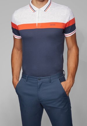 Homme portant un polo rayé blanc, orange et bleu marine avec un pantalon bleu, les mains dans les poches, devant un fond gris.