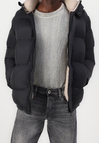 Chaqueta de plumas negra con un forro beige, combinada con un suéter de canalé gris claro y jeans gris oscuro. La chaqueta tiene cierre de cremallera y capucha.