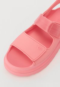 Rosa gummisandaler med borrelåsremmer, teksturert fotseng og UGG-logo. Lukket tå og åpen bakdesign, med en tykk såle.