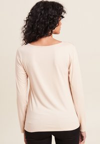 Langärmliges, eng anliegendes beiges Oberteil mit Rundhalsausschnitt und einer glatten, weichen Textur; hat einen abgerundeten Saum auf Taillenhöhe und minimale Nähdetails.