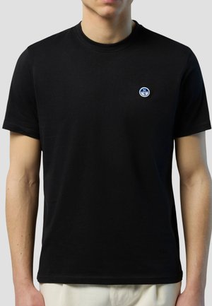 Uomo che indossa una maglietta nera a maniche corte con un piccolo logo rotondo blu e bianco sul lato sinistro del petto, in piedi contro uno sfondo uniforme.