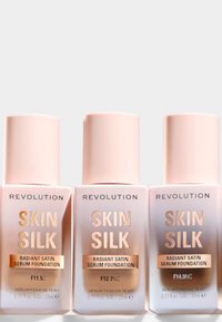 Tre flasker med Revolution Skin Silk Radiant Satin Serum Foundation. Flaskene er lys rosa med gulltext. Hver har forskjellige fargeidentifikatorer.