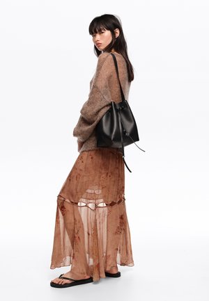 Mujer con cabello largo y oscuro que lleva un suéter marrón peludo, falda maxi floral translúcida, chanclas negras y lleva un bolso de cuero negro tipo cubo.