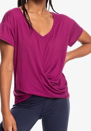 Osoba ubrana w magentowy, krótki rękaw, top z dekoltem w serek z marszczonym przodem oraz ciemnoniebieskie dopasowane spodnie, pozująca z ręką na biodrze.