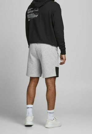 Personne portant un sweat à capuche noir avec du texte blanc dans le dos, un short gris clair avec une tache foncée, des chaussettes blanches et des baskets blanches, se tenant sur un sol gris.