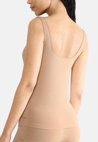 Beige nauwsluitende tanktop met brede schouderbanden, een gladde textuur, een lage rugontwerp en subtiele branding nabij de zoom.