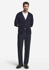 Marinblå stickad cardigan med v-ringning och fem knappar, kombinerad med en vit t-shirt och mörka skräddarsydda byxor. Svarta skor fulländar stilen.