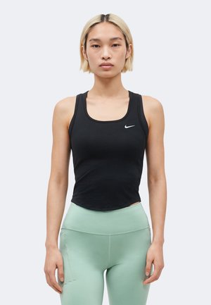 Femme aux cheveux blonds courts portant un débardeur noir Nike et des leggings taille haute vert clair, debout devant un fond uni.