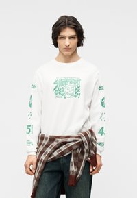 Νέος μεσαίου μήκους σκούρα μαλλιά που φοράει ένα λευκό μπλουζάκι με μακριά μανίκια και ένα καφέ καρό πουλόβερ δεμένο γύρω από τη μέση.