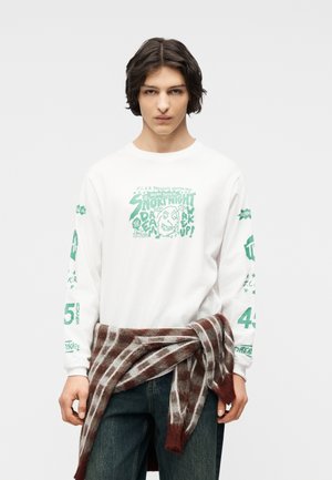 Jeune personne aux cheveux bruns mi-longs portant un t-shirt blanc à manches longues avec un motif graphique et un pull à carreaux marron noué autour de la taille.