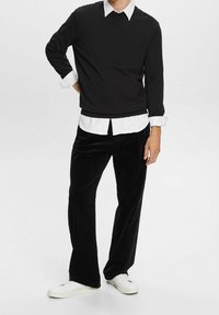 Musta huppari pyöreällä kaula-aukolla valkoisen nappipaidan päällä, yhdistettynä mustiin samettihousuihin ja valkoisiin tennareihin.