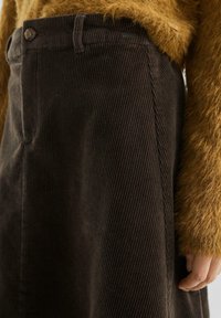 Bruine corduroy rok gedragen met een fluffige mosterdkleurige trui, waarbij de taille, knoop, riemlussen en een hand naast de rok zichtbaar zijn.