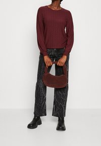 Bordeauxfarbener, strukturierter Langarmtop, kombiniert mit schwarzen, gemusterten Hosen. Das Model hält eine kleine braune Tasche aus Kunstfell. Schwarze Ankle Boots runden den Look ab.