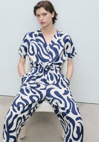 Blue and white patterned jumpsuit with short sleeves, button-up front, and a tied waist. Soft fabric with a relaxed fit and side pockets.