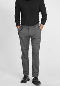 Homme portant un pantalon gris sur mesure, une chemise noire à manches longues, des chaussures en cuir noires, les mains dans les poches, sur un fond uni.