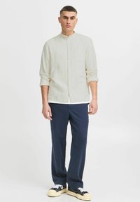 Joven de pie con una camisa abotonada color blanco roto y mangas arremangadas, pantalones azul marino y zapatillas negras y blancas, sobre un fondo blanco.
