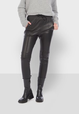 Persoon die zwarte leren broek draagt met elastische taille en trekkoord, gecombineerd met zwarte enkellaarzen en een lichtgrijze top met lange mouwen.
