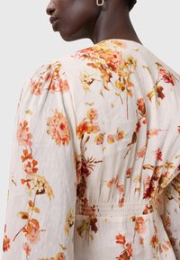 Tessuto leggero di colore crema con stampa floreale in rosa, arancione e verde. Presenta maniche a sbuffo e un dettaglio arricciato in vita.