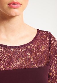 Burgundské tričko s krajkovým potahem, který obsahuje složité vzory vlnění, kombinované s pevným materiálem pod ním. Klasický kulatý výstřih a strukturovaný povrch.