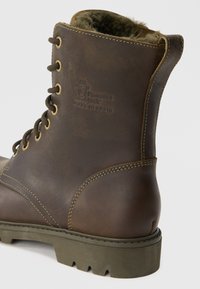 Bota de caminhada de couro castanho com acabamento texturizado, colarinho almofadado, sola de borracha verde e costura amarela em contraste. Logótipo em relevo na lateral.