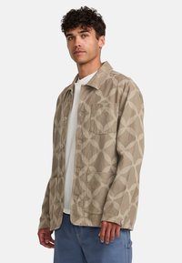 Beige patterned jacket with a lightweight fabric, featuring a collar, front buttons, and a pocket on the left chest.