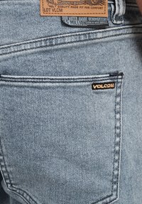 Vaqueros de mezclilla azul claro con un acabado texturizado, presentando un bolsillo trasero con un pequeño logo negro "VOLCOM" y una etiqueta de cuero de la marca.