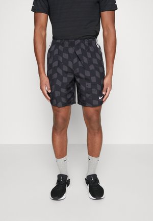 Sportowiec ubrany w czarne, wzorzyste krótkie spodenki sportowe, czarną koszulkę, szare skarpetki marki Nike oraz czarne buty do biegania Nike, stoi na jednolitym tle.